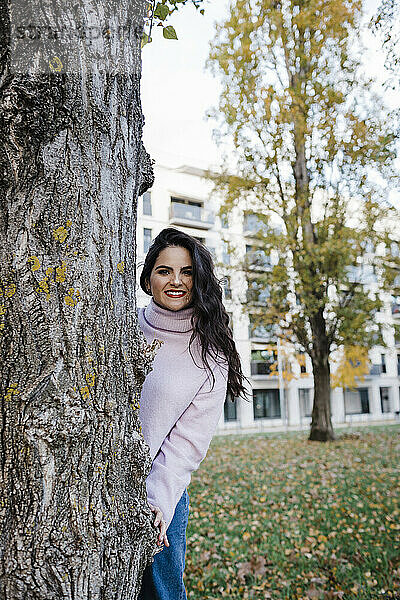 Lächelnde junge schöne Frau  die den Herbst genießt und hinter einem Baumstamm im Park steht