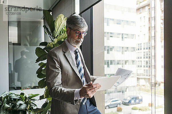 Geschäftsmann mit Dokumenten unter Verwendung eines Tablet-PCs  der am Fenster im Büro steht