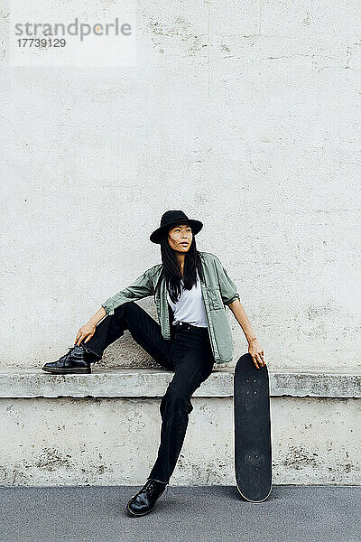 Junge Frau mit Hut sitzt mit Skateboard an der Wand