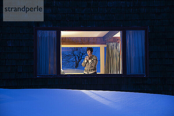 Frau schaut in der Abenddämmerung im Winter durch das Fenster