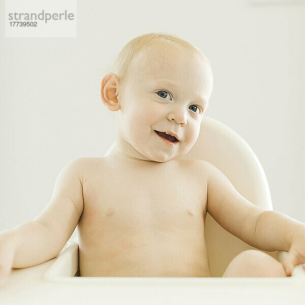 Porträt eines hemdlosen Jungen (12–17 Monate)  der im Hochstuhl sitzt