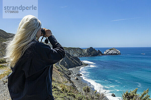 USA  Kalifornien  Big Sur  Rückansicht einer älteren blonden Frau  die mit einem Fernglas auf das Meer an der Küste von Big Sur blickt