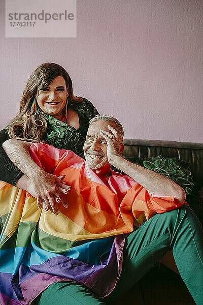Glücklicher Transgender mit schwulem Mann auf Sofa sitzend gegen rosa Wand im Wohnzimmer