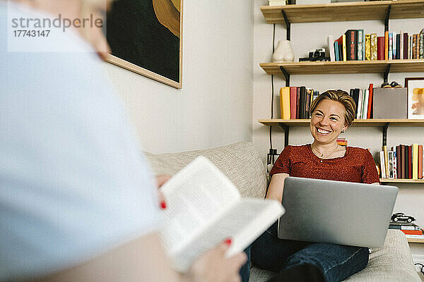 Lächelnde lesbische Frau  die freiberuflich am Laptop arbeitet  während sie mit ihrer Freundin im Wohnzimmer spricht