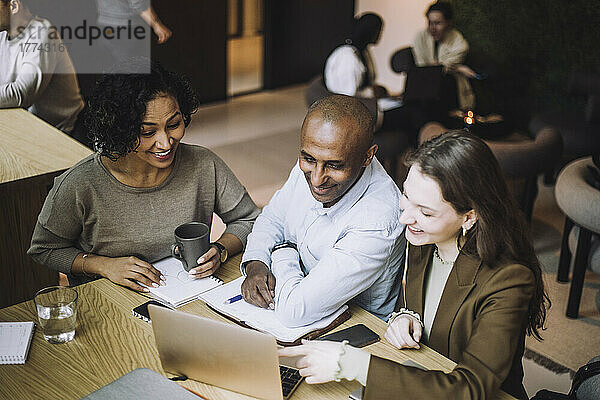 Lächelnde männliche und weibliche Kollegen teilen sich einen Laptop  während sie am Schreibtisch in einem kreativen Büro sitzen und diskutieren