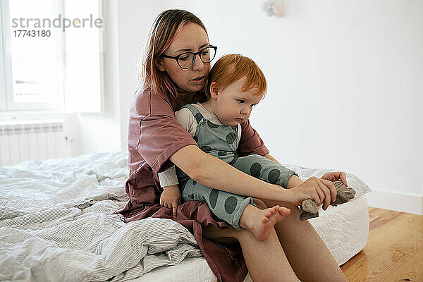 Mutter zieht männlichem Kleinkind  das im Schlafzimmer sitzt  eine Socke über den Fuß
