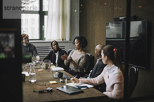 Männliche und weibliche Kollegen betrachten eine Geschäftsfrau  die am Konferenztisch sitzend Ideen austauscht  während sie gestikulieren