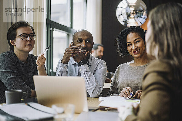 Lächelnde männliche und weibliche Kollegen betrachten eine Geschäftsfrau  die am Schreibtisch in einem kreativen Büro einen Plan bespricht