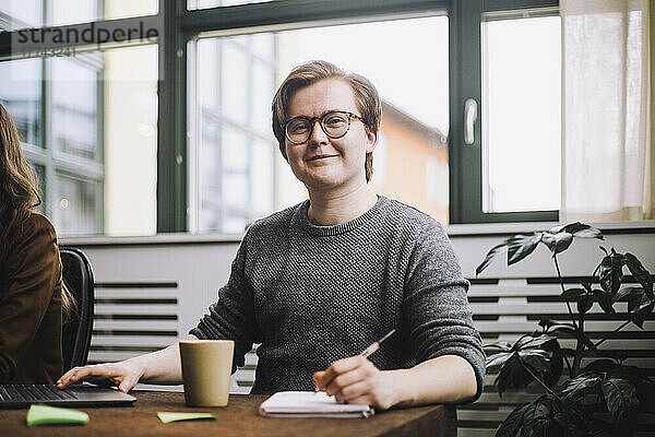 Porträt eines lächelnden jungen Geschäftsmannes mit Brille  der mit einem Tagebuch am Konferenztisch sitzt