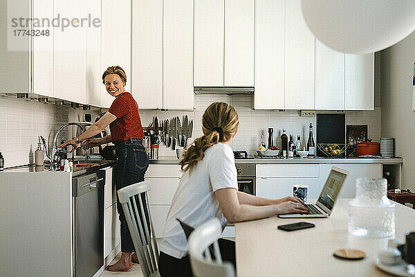 Freiberufler  der am Laptop arbeitet  während er mit seiner Freundin spricht  die zu Hause in der Küche Hausarbeiten erledigt