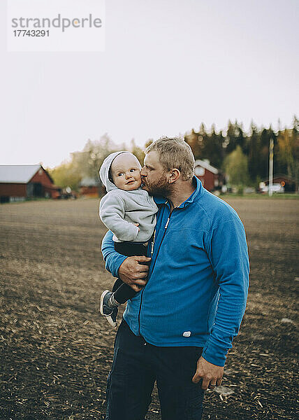 Vater küsst Kleinkind Sohn stehen auf dem Bauernhof bei Sonnenuntergang