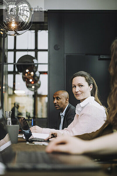 Porträt einer Geschäftsfrau im mittleren Erwachsenenalter  die mit einem Laptop neben Kollegen in einem Besprechungsraum sitzt