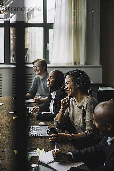 Lächelnde Unternehmerin mit Hand am Kinn  die mit Kollegen am Konferenztisch sitzt