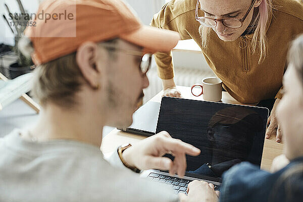 Nicht-binärer Computerprogrammierer  der auf den Laptop-Bildschirm schaut  während Kollegen bei einer Besprechung im Büro diskutieren