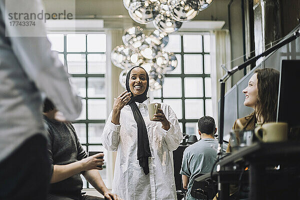 Lächelnde junge Geschäftsfrau  die eine Kaffeetasse hält  während sie mit Kollegen in einem kreativen Büro Ideen austauscht