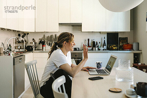 Glückliche Freiberuflerin winkt bei einem Videoanruf über den Laptop am Küchentisch