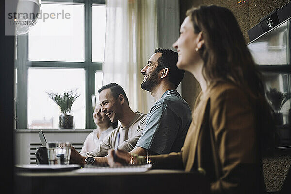 Glückliche Geschäftsleute sitzen mit Kollegen am Konferenztisch in einem kreativen Büro