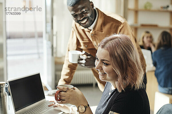Seitenansicht einer lächelnden Unternehmerin  die mit einem Kollegen im Büro eine Kaffeetasse hält