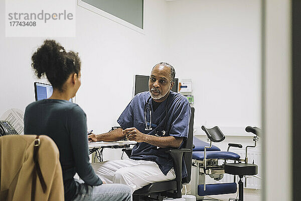 Reifer männlicher Arzt im Gespräch mit einer Patientin  die am Schreibtisch in einer medizinischen Klinik sitzt