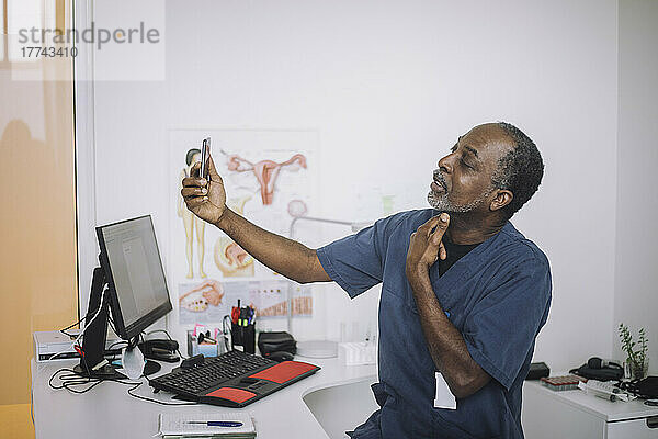 Männlicher HNO-Arzt bei einer Online-Konsultation per Videoanruf auf einem Smartphone  während er am Schreibtisch in der Klinik sitzt
