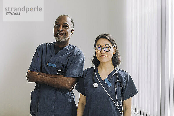 Porträt eines Arztes  der mit verschränkten Armen neben einer Kollegin aus dem Gesundheitswesen an einer weißen Wand im Krankenhaus steht