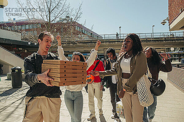 Mann trägt einen Stapel Pizzakartons  während er mit Freunden an einem sonnigen Tag auf der Straße spazieren geht