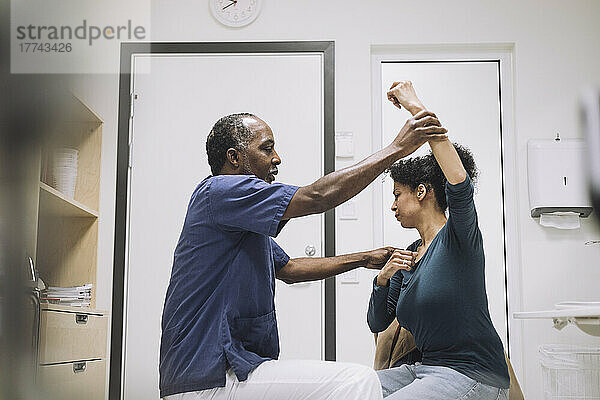 Seitenansicht eines männlichen Arztes  der einer weiblichen Patientin im Krankenhaus sitzend mit erhobener Hand eine physikalische Therapie gibt