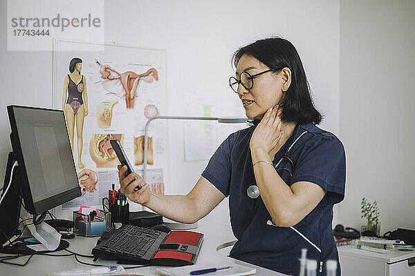 Weibliche HNO-Ärztin bei einem Videoanruf über ein Smartphone  die eine Online-Konsultation durchführt  während sie in der Klinik den Hals erklärt