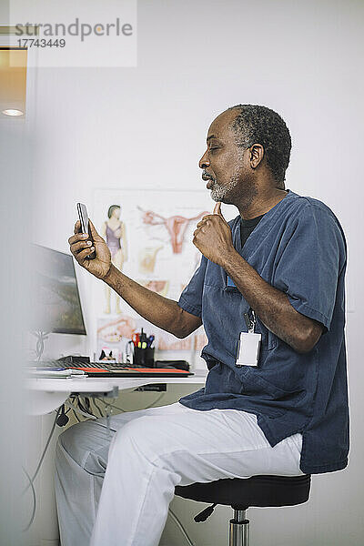 Männlicher Arzt bei der Online-Konsultation über das Mobiltelefon  während er am Schreibtisch in der Klinik sitzt