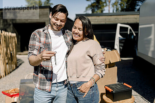 Lächelndes Paar betrachtet den Schlüssel des neuen Hauses  während es vor dem neuen Haus steht