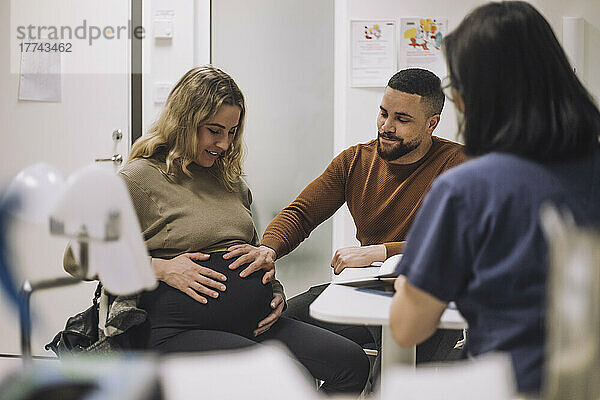 Lächelnder Mann  der den Bauch einer schwangeren Frau berührt  während er mit einem Gynäkologen in einer medizinischen Klinik spricht
