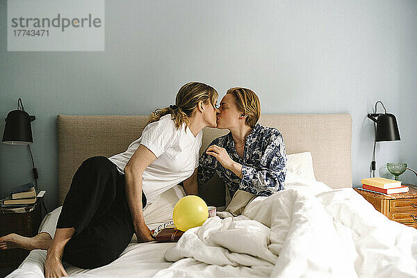 Lesbische Frau mit Geburtstagsgeschenk küsst Freundin auf dem Bett zu Hause