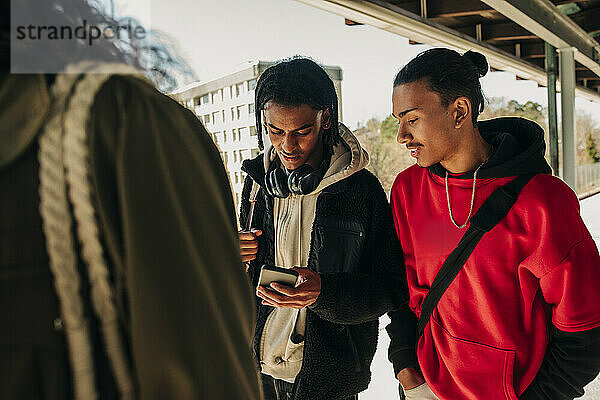 Junger Mann teilt sein Smartphone mit einem Freund  während er am Bahnsteig spazieren geht