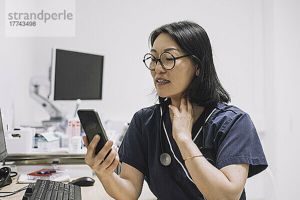 Eine HNO-Ärztin erklärt  während sie den Hals während einer Online-Konsultation über ein Smartphone in einer medizinischen Klinik berührt.