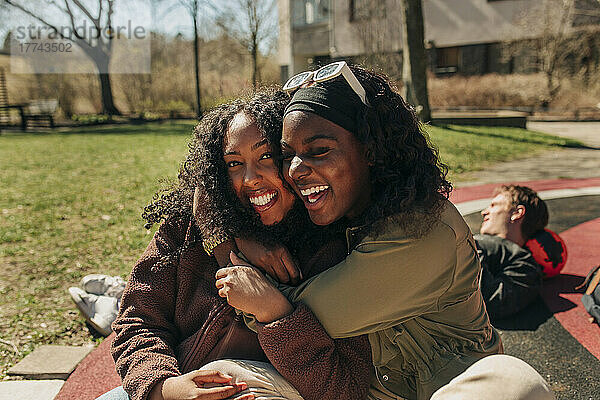 Glückliche multirassische Freundinnen  die sich gegenseitig umarmen  während sie auf einem Spielplatz an einem sonnigen Tag sitzen
