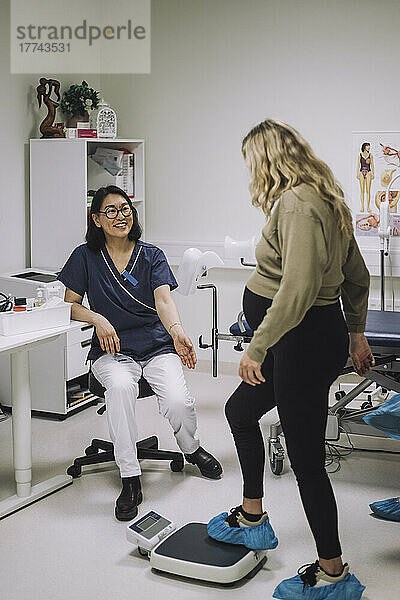 Lächelnde Ärztin in voller Länge  die eine schwangere Frau auf der Waage in einer medizinischen Klinik betrachtet