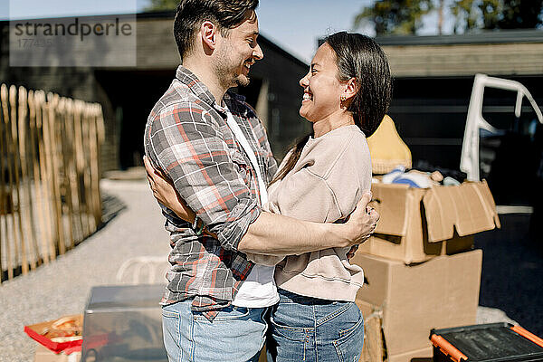 Glückliches Paar  das sich umarmt  während es an einem sonnigen Tag vor einem neuen Haus steht