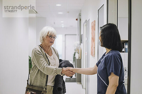 Lächelnde ältere Frau schüttelt Hände mit einer Ärztin  die an der Tür eines Krankenhauses steht