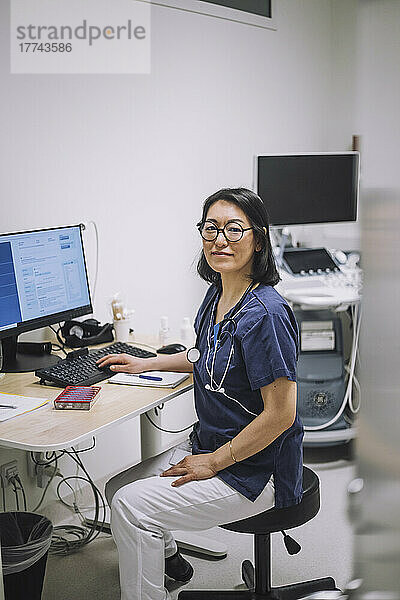 Porträt einer lächelnden Ärztin mit Brille  die am Schreibtisch in einer medizinischen Klinik sitzt