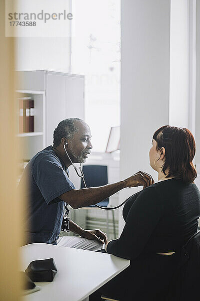 Reifer männlicher Arzt mit Stethoskop  der eine Patientin am Schreibtisch sitzend im Krankenhaus untersucht