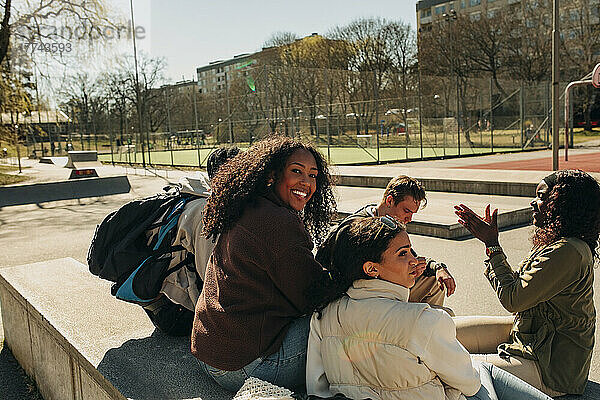 Lächelndes Mädchen  das über die Schulter schaut  während es mit Freunden auf einer Stützmauer im Park sitzt