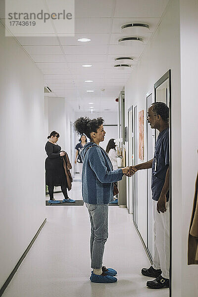 Seitenansicht eines männlichen Gesundheitsexperten  der einer Patientin im Korridor die Hand schüttelt