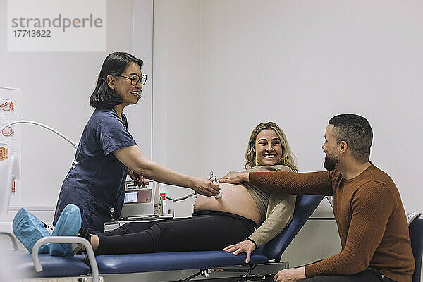 Glücklicher Gynäkologe bei der Ultraschalluntersuchung mit Blick auf einen Mann  der den Bauch einer schwangeren Frau im Krankenhaus berührt