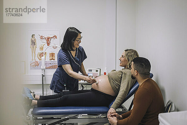 Lächelnde Ärztin bei der Ultraschalluntersuchung einer schwangeren Frau auf dem Untersuchungstisch eines Mannes im Krankenhaus