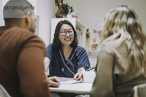 Glückliche weibliche Gesundheitsexpertin  die mit einem Paar in einer medizinischen Klinik über ein Krankenblatt diskutiert