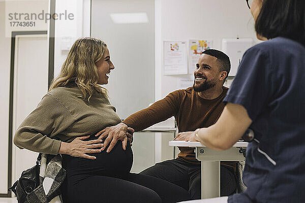 Glücklicher Mann  der den Bauch einer schwangeren Frau berührt  während er mit einer Ärztin in einer medizinischen Klinik sitzt