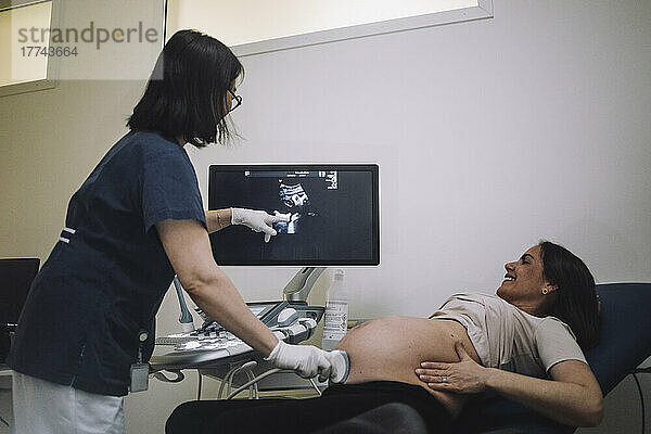 Weiblicher Gynäkologe  der auf den Bildschirm zeigt  während er eine schwangere Frau in einer medizinischen Klinik mit Ultraschall untersucht