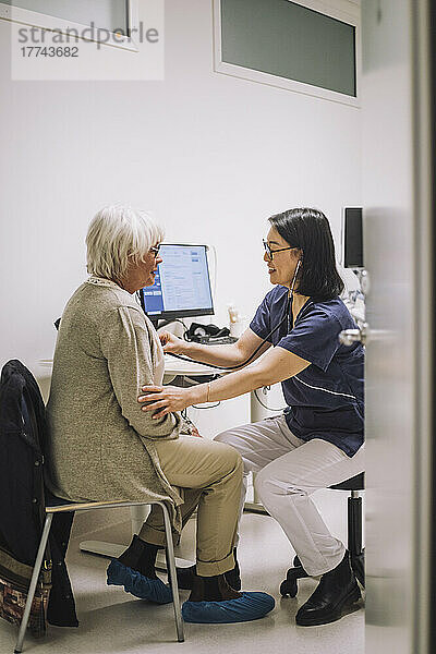 Weiblicher Gesundheitsexperte mit Stethoskop in voller Länge bei der Untersuchung eines älteren Patienten durch die Tür in einer medizinischen Klinik