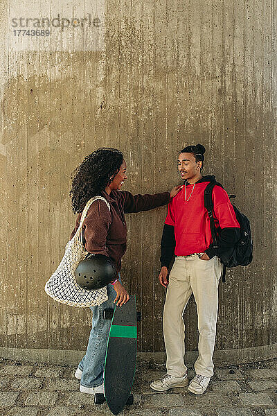 Mann mit Hand in der Tasche im Gespräch mit einer Freundin  die ein Skateboard hält  während er an der Wand steht