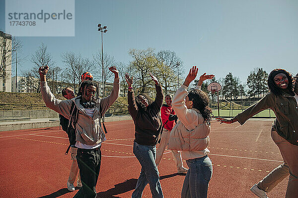 Unbeschwerte multirassische Freunde mit erhobenen Armen tanzen zusammen auf dem Sportplatz an einem sonnigen Tag